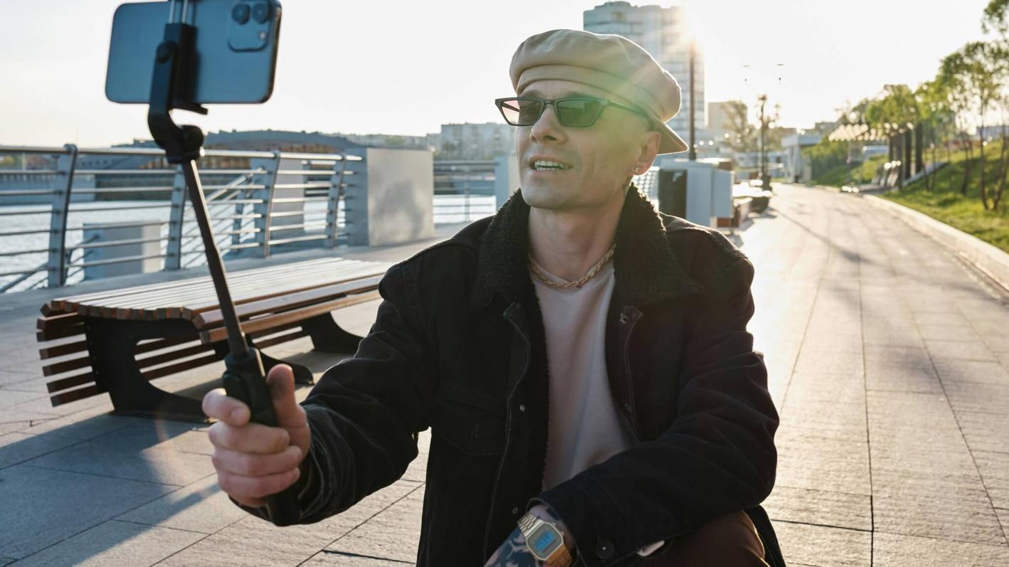 Man in a hat and shades holding a smartphone in a selfie stick