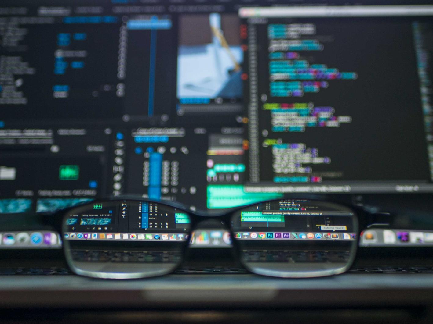 A photo of a pair of glasses in focus, with monitors showing code, blurred in the background